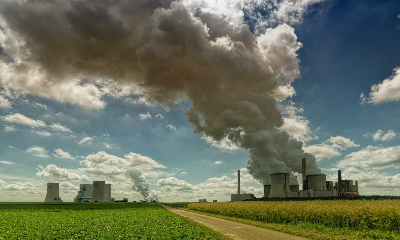 Power plant in rural landscape emitting smoke under a bright blue sky with fluffy clouds.