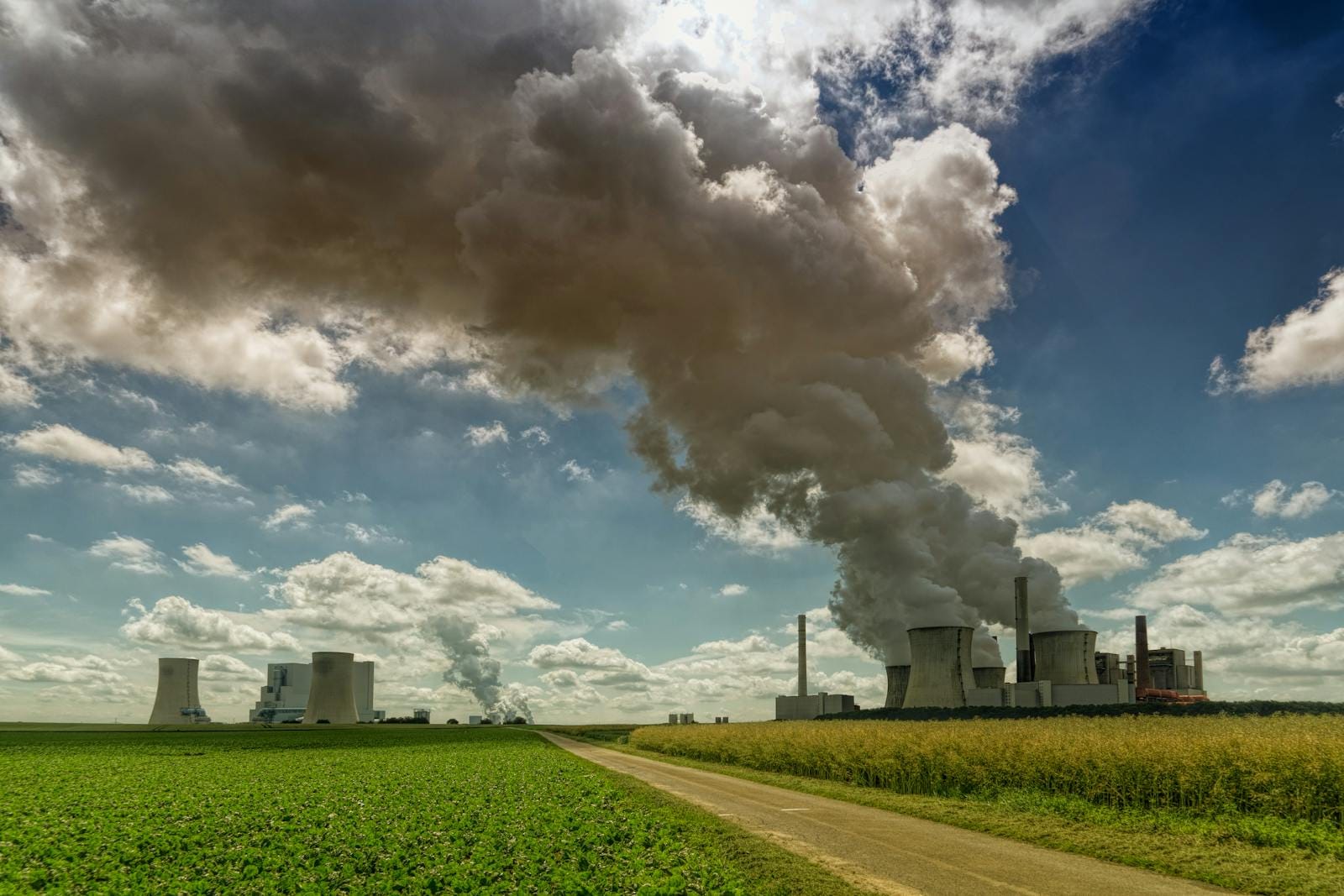 Power plant in rural landscape emitting smoke under a bright blue sky with fluffy clouds.